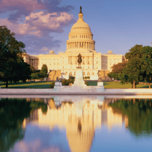The U.S. Capitol building at sunset, with its dome illuminated, reflected in a calm pool in the foreground, surrounded by green trees and a statue in front.