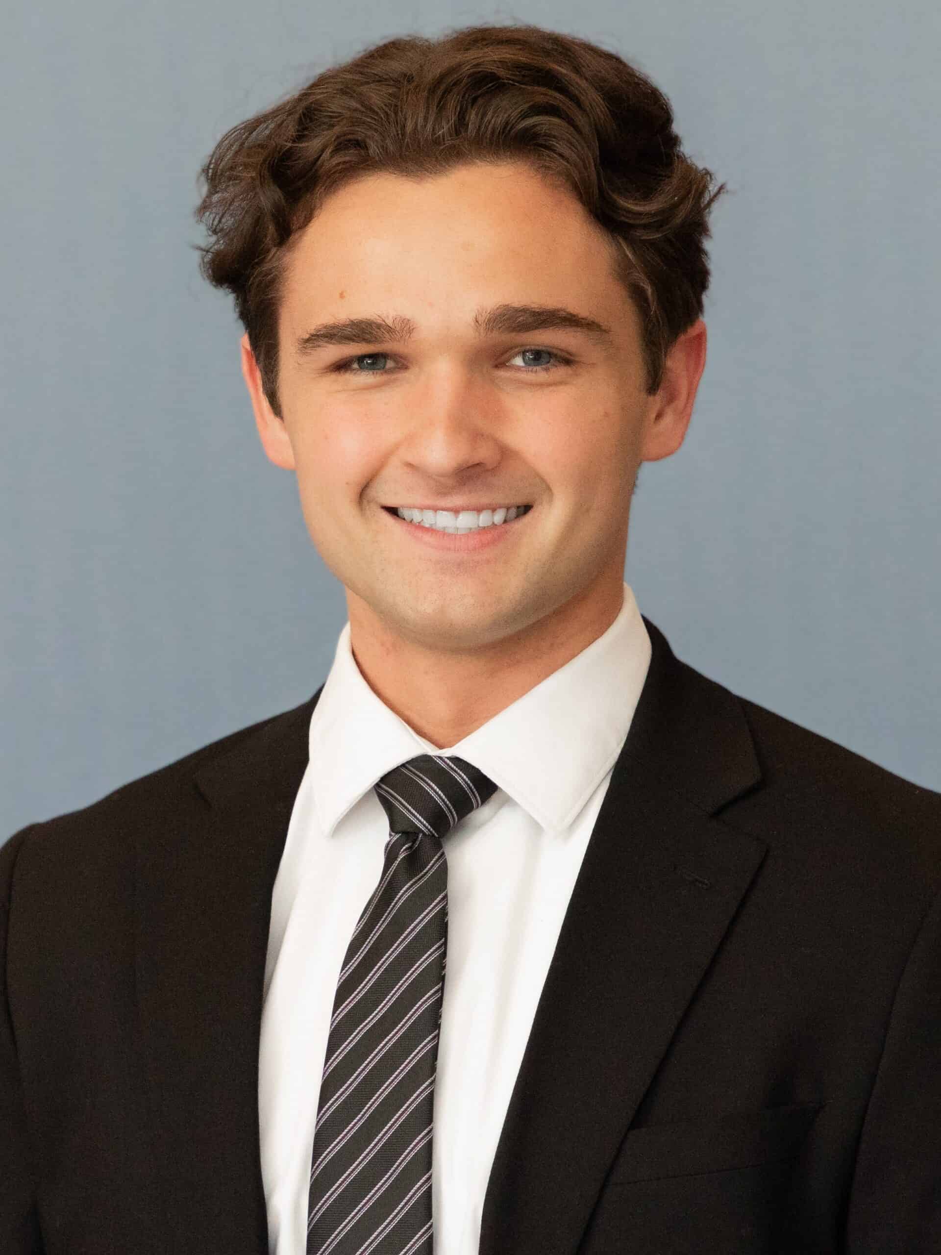 A young man with wavy brown hair is smiling, wearing a black suit, white dress shirt, and striped tie, standing against a plain light blue background—perfectly dressed for making Capital Connections.