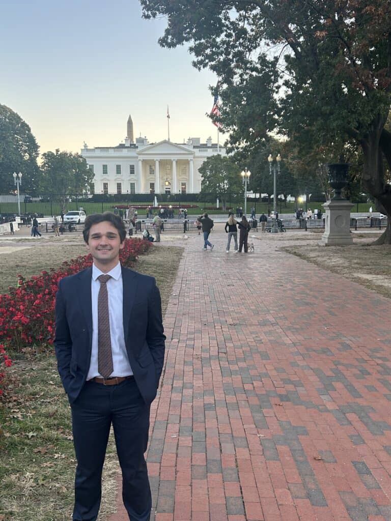 A man in a suit stands on a brick walkway with his hands in his pockets, smiling in front of the White House—an image that captures the essence of Capital Connections amidst people and trees in the background.