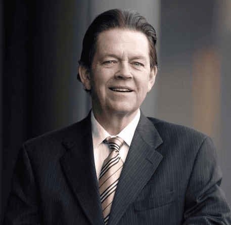 A middle-aged man in a dark pinstripe suit, white shirt, and striped tie stands indoors, smiling confidently with a neutral background.