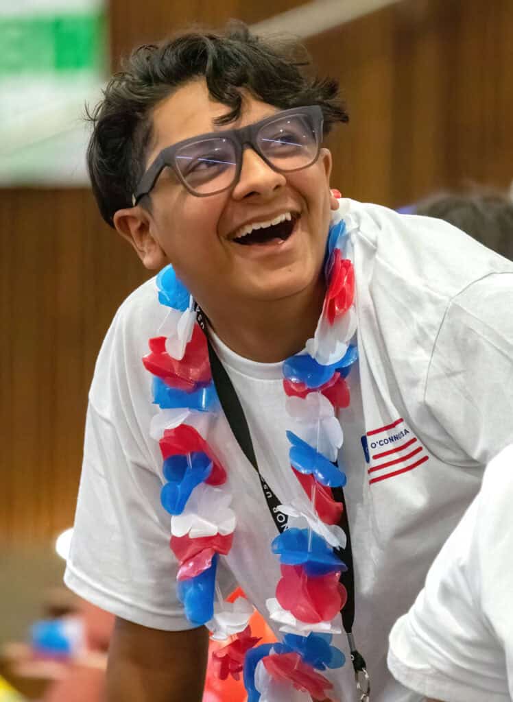 A smiling person wearing glasses, a white shirt, and a red, white, and blue lei with a name tag and lanyard laughs while leaning forward indoors at The Citizen.