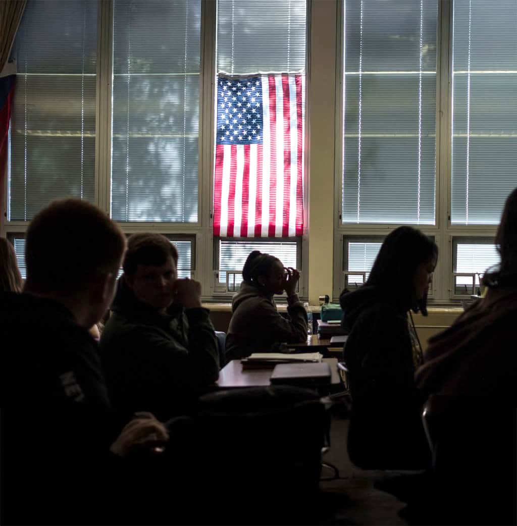 Students sit at desks in a dimly lit classroom with large windows. Sunlight filters through blinds and an American flag, which hangs in the center window, creating a silhouette effect—a fitting scene for teaching civics.