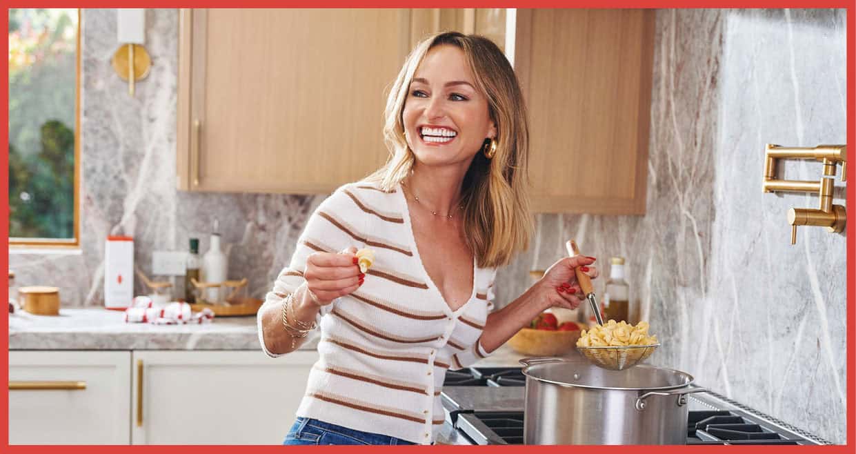 A woman, embodying the spirit of women in leadership, stands in her kitchen with a pasta shell in one hand and a strainer of pasta in the other. Smiling beside the stove, she reflects a legacy luncheon atmosphere amid wooden cabinets and a marble backsplash.