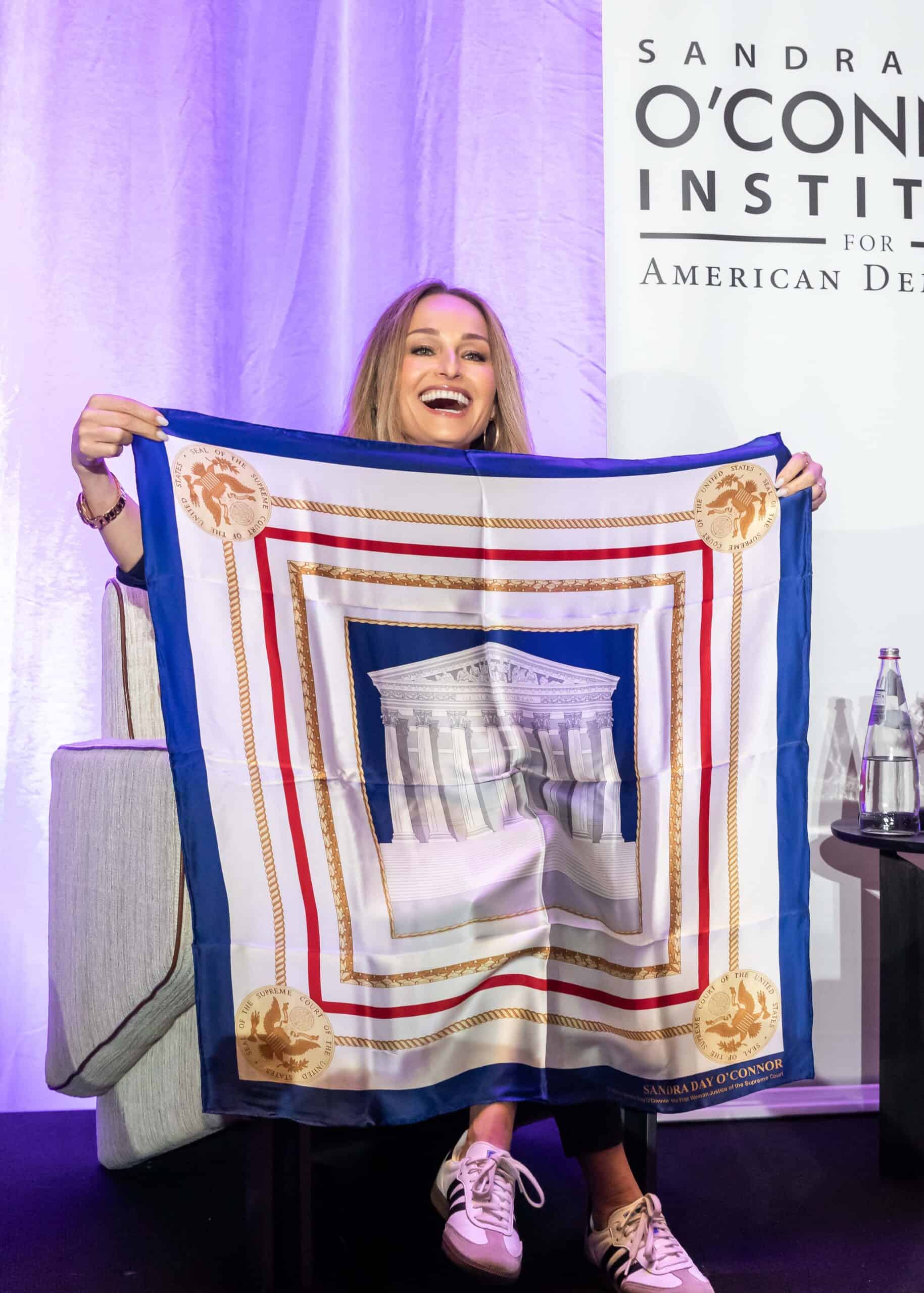 A smiling woman sits on a chair, holding up a large decorative scarf featuring an illustration of a neoclassical building, possibly a courthouse, with gold circular emblems in the corners. A Sandra Day O’Connor Institute sign is behind her.