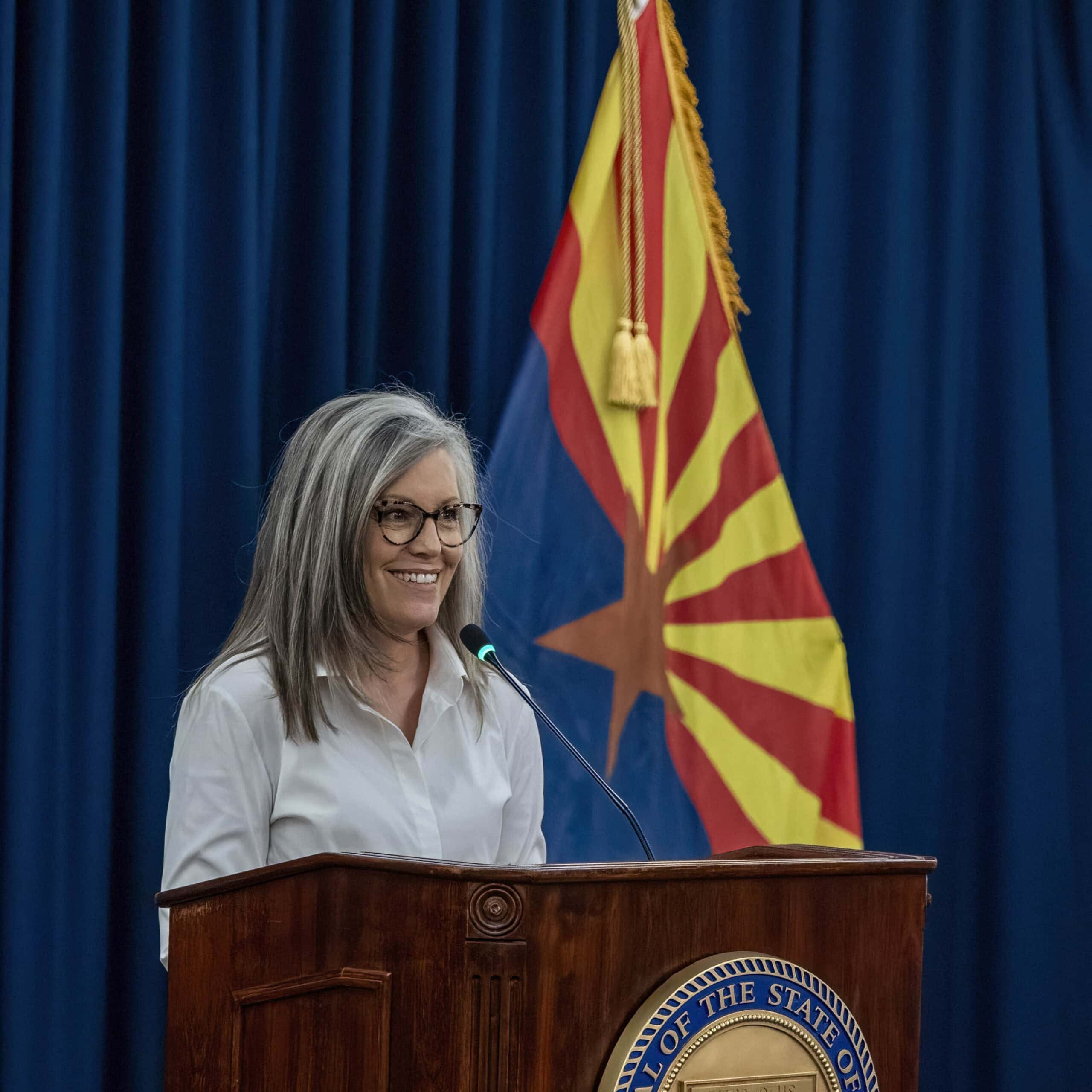 A gray-haired woman with glasses stands at a podium, smiling and speaking into a microphone. Behind her is a blue curtain and the Arizona state flag, highlighting her role as an active citizen in the community.