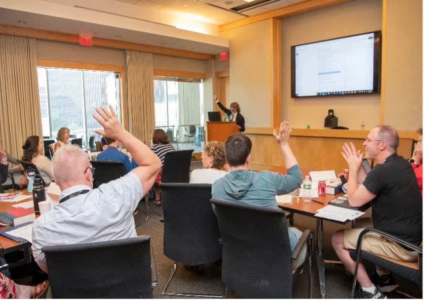 Several people seated around a conference table raise their hands during a meeting or presentation. A person stands at a podium, and a large screen displays information about the Constitution at the front of the room.