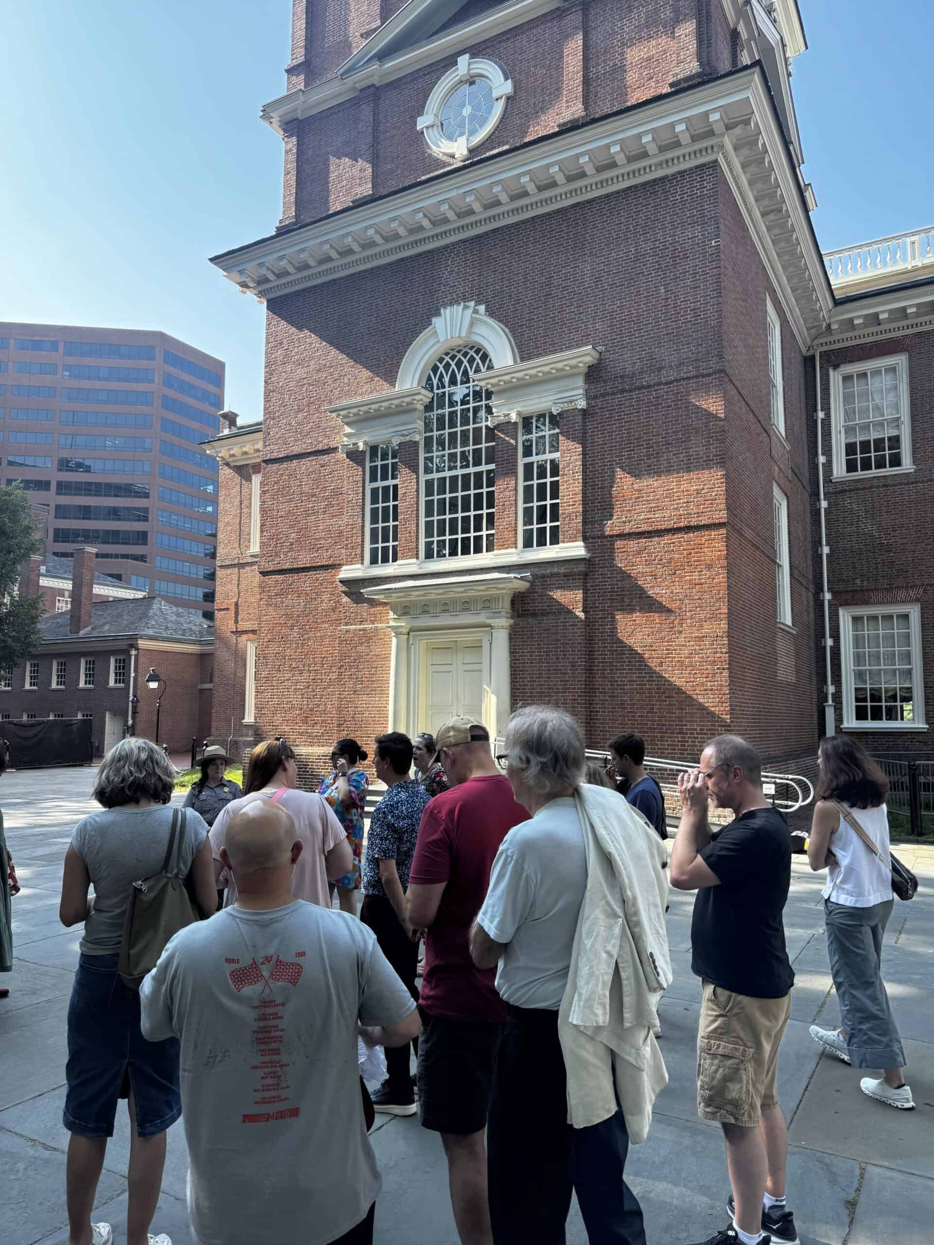A group of people stand outside a historic red brick building with tall windows, listening to a tour guide discuss the Constitution on a sunny day. A modern office building is visible in the background.