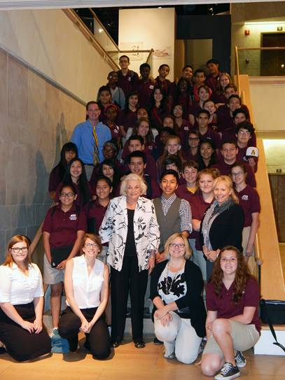 A large group of students in maroon polo shirts and khaki pants poses on a staircase, with several adults standing and sitting at the front, all smiling at the camera indoors—celebrating the spirit of the O'Connor Legacy.