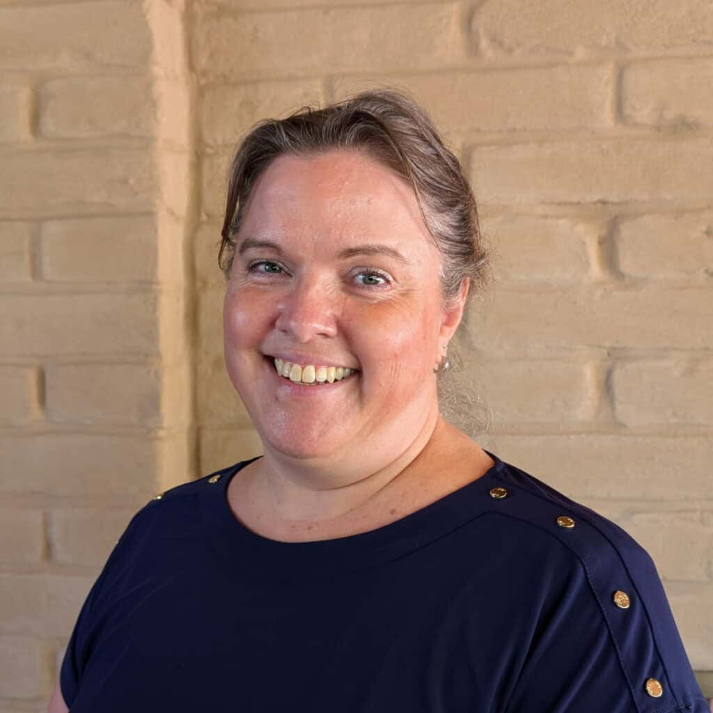A woman with light skin and brown hair pulled back smiles at the camera. She is wearing a navy blue top with gold buttons and stands in front of a beige brick wall.