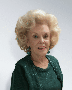 An elderly woman with styled blonde hair, wearing a sparkly green top and green earrings, smiles at the camera against a plain white background.
