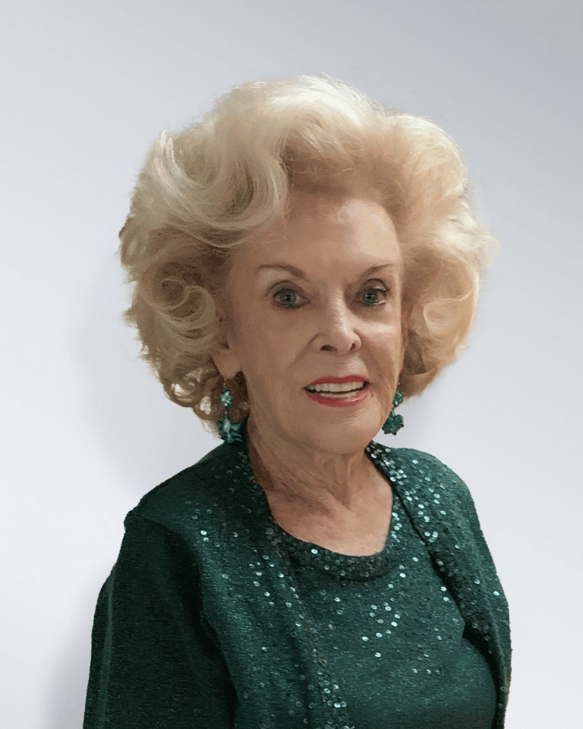 An elderly woman with styled blonde hair, wearing a sparkly green top and green earrings, smiles at the camera against a plain white background.