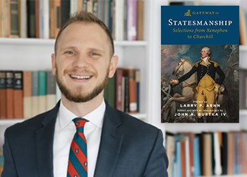 A smiling person in a suit and tie stands in a library. A book titled "Statesmanship: Selections from Xenophon to Churchill" by Larry P. Arnn is visible, featuring an image of an 18th-century military officer. Bookshelves are in the background.