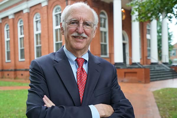 An older man with white hair and a mustache stands confidently with arms crossed in front of a brick building with columns. He is wearing a dark suit, light blue shirt, and red patterned tie. The background shows a lawn and pathway.