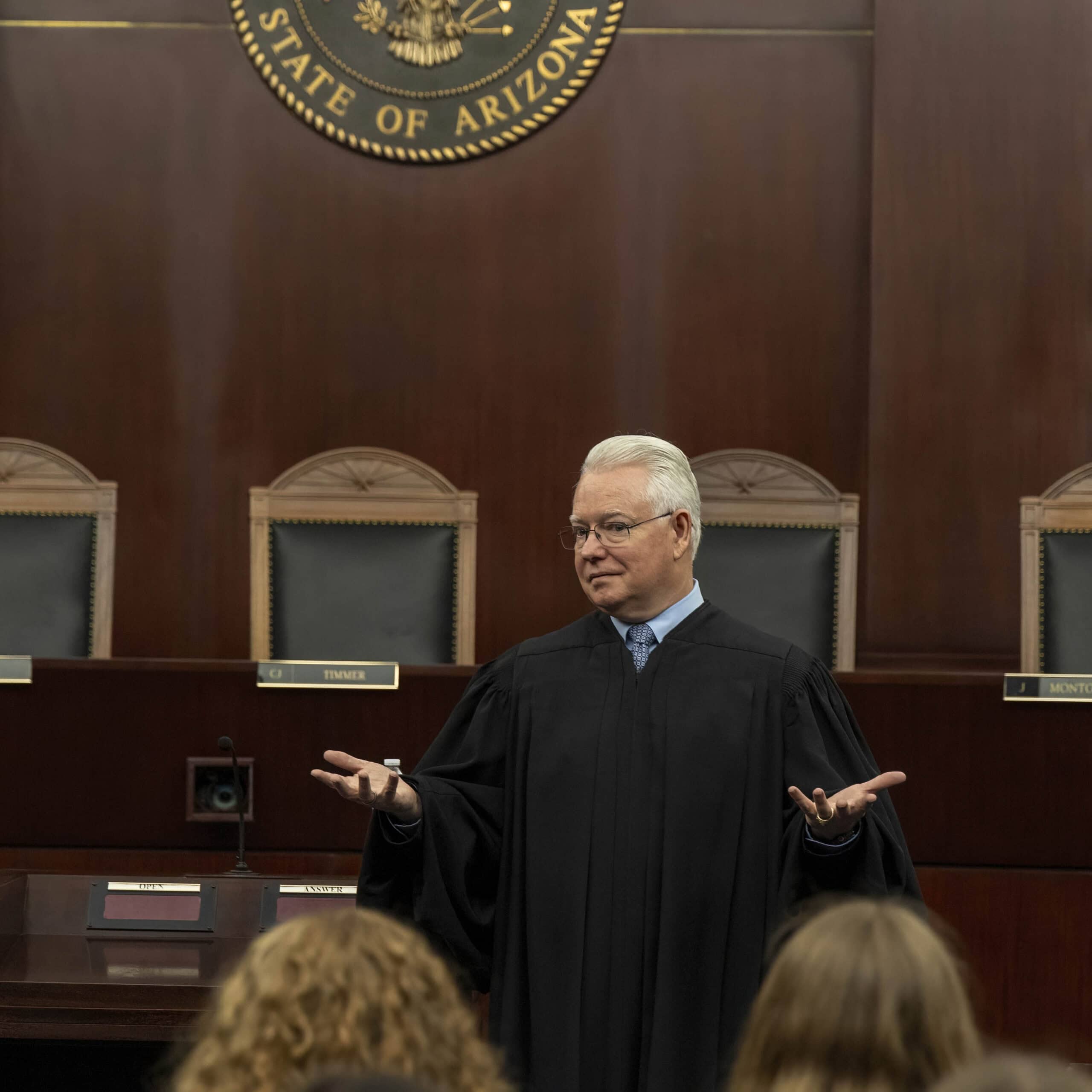 A judge in a black robe stands before empty chairs and a wooden bench in a courtroom, gesturing with open hands while addressing citizens seated in the foreground. The Arizona state seal is visible behind him.
