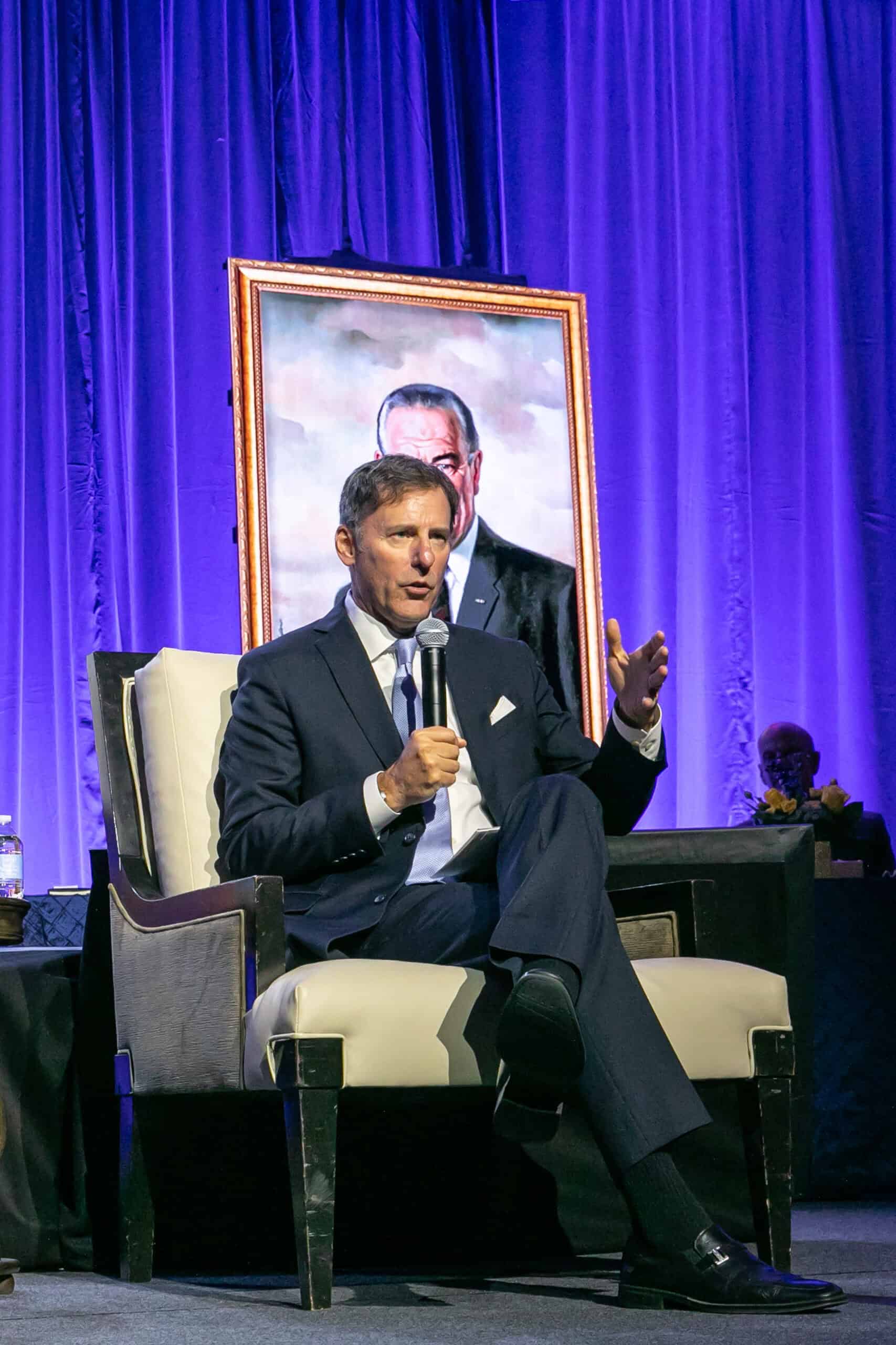 A man in a dark suit sits in a chair holding a microphone, speaking on stage before a large portrait of another man and a blue-purple curtain, reminiscent of events featuring historians like Doris Kearns Goodwin.