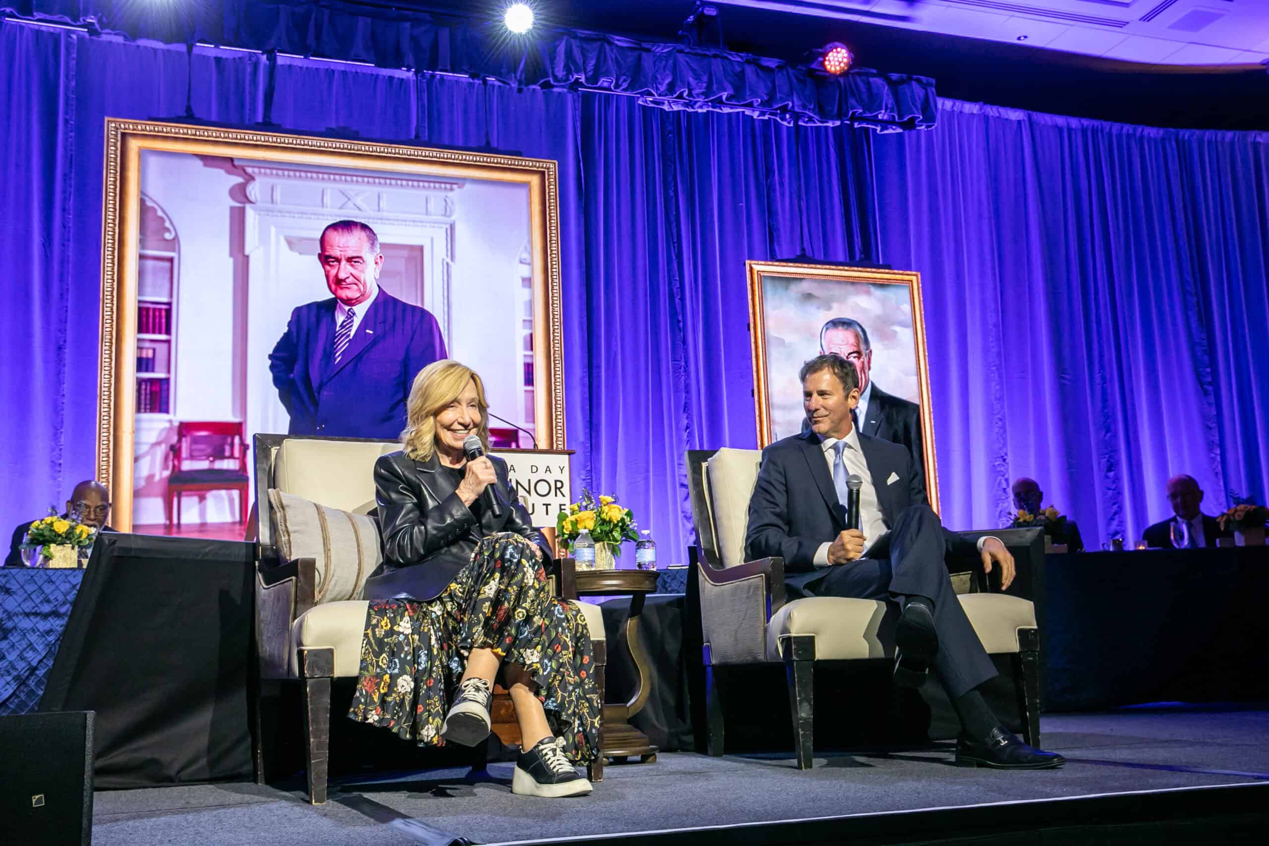 A woman and a man, including Doris Kearns Goodwin, sit on stage in armchairs, smiling and speaking into microphones. Behind them are two large portraits and a purple curtain, with flowers decorating the stage and people seated at a table in the background.