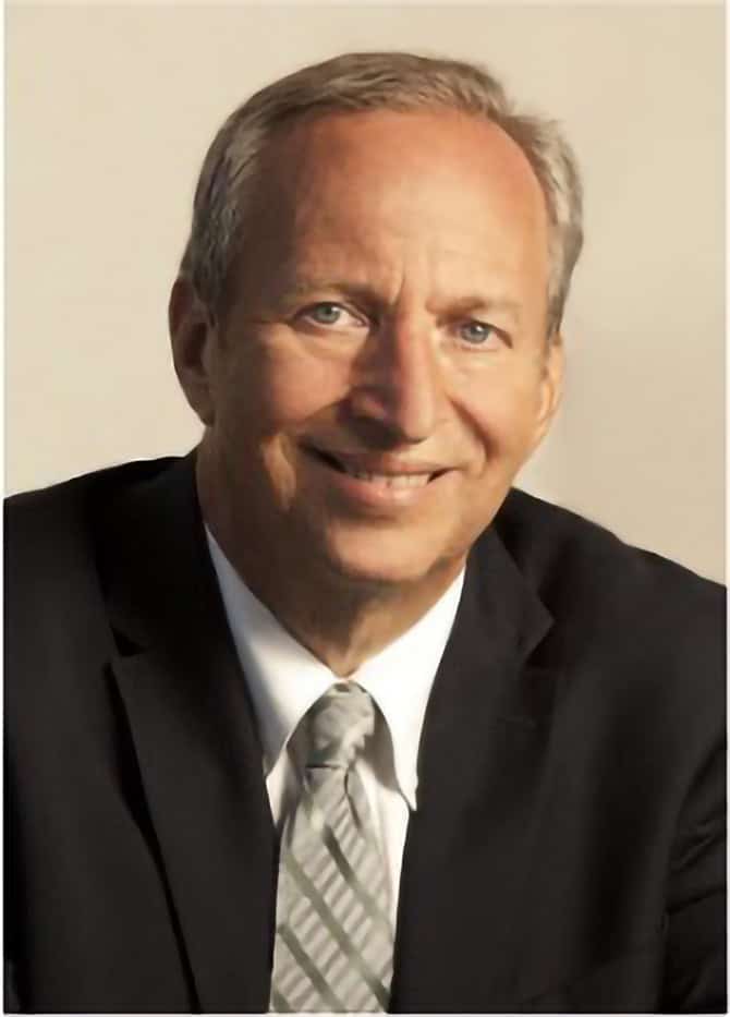 A middle-aged man with short gray hair, wearing a black suit, white shirt, and striped tie, smiles at the camera against a plain light background, presenting the professional image of a 501(c)(3) organization’s executive.