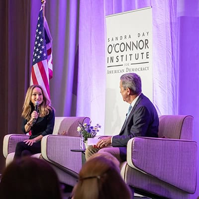 A woman and a man sit on stage in armchairs, engaged in conversation at the Sandra Day O’Connor Institute for American Democracy event. An American flag and purple lighting are visible in the background.