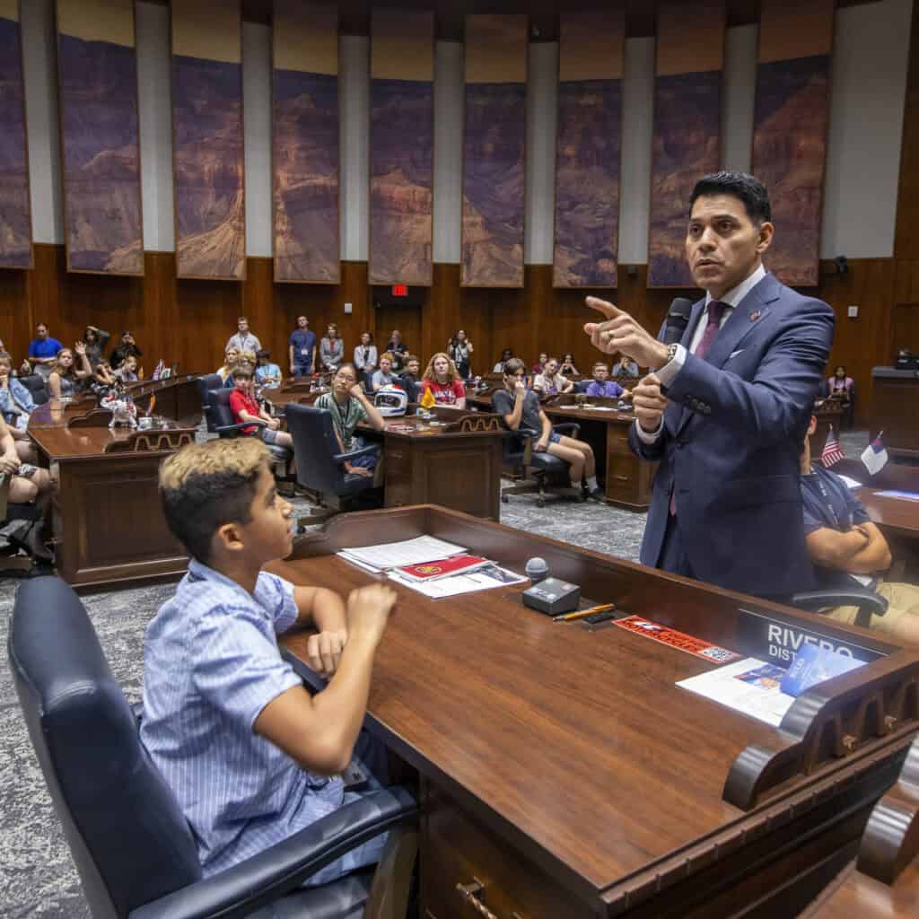 A man in a suit speaks to a seated boy in a government chamber filled with people. The audience listens attentively, creating a formal and educational atmosphere focused on building thoughtful citizens.