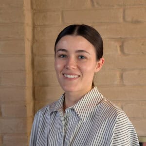 Lily Oswald, a woman with dark hair pulled back, smiles at the camera. She wears a white shirt with thin black stripes and stands in front of a tan brick wall.