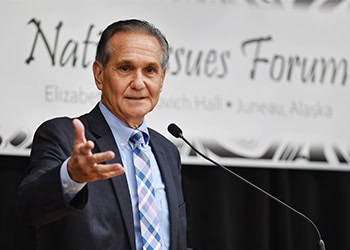 A man in a suit speaks at a podium with a microphone. A sign behind him partially reads "National Issues Forum" and mentions "Juneau, Alaska." He gestures with his left hand while speaking.