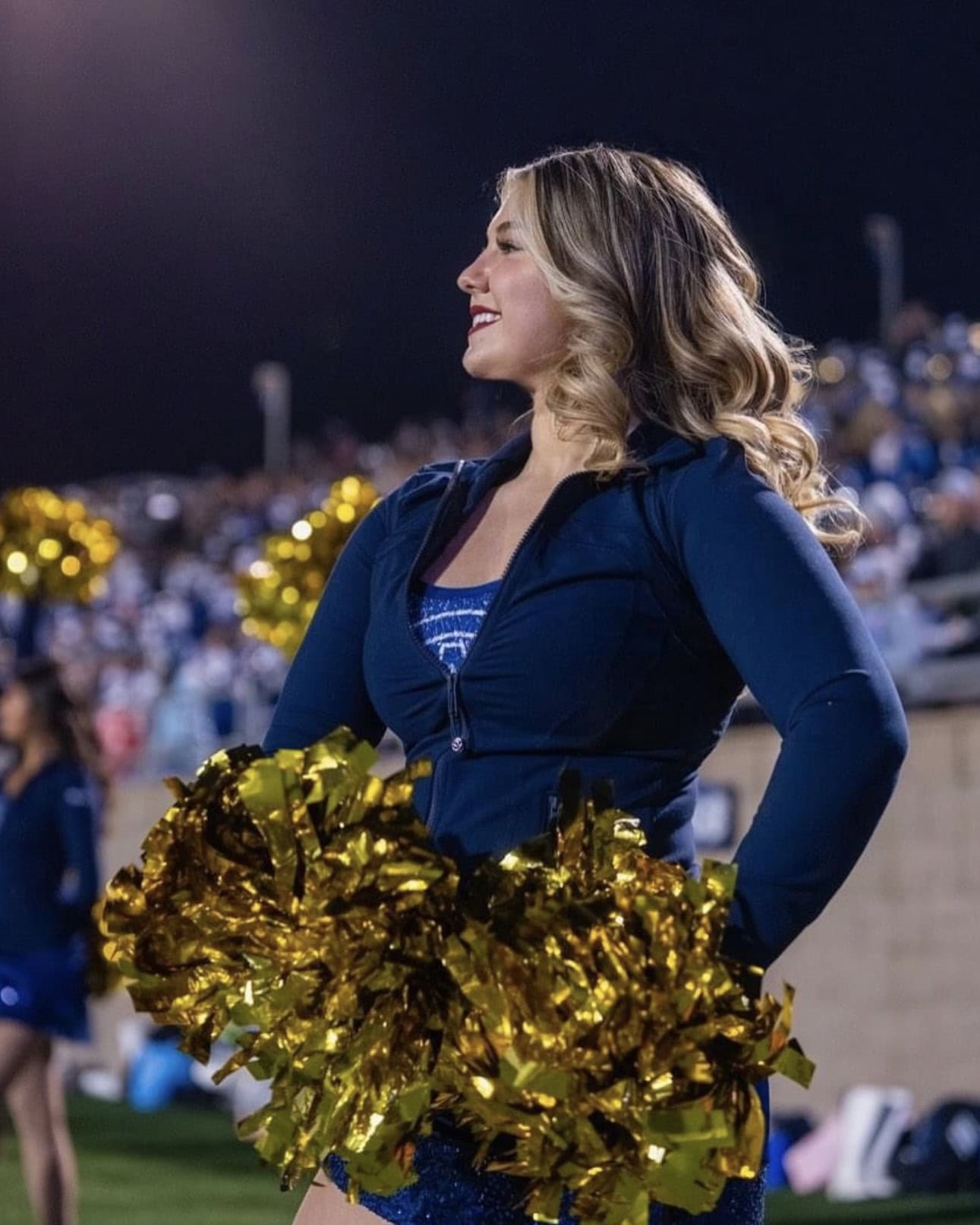 A cheerleader with wavy blonde hair, wearing a navy blue outfit and holding gold pom-poms, smiles while standing on a field at night with a blurred crowd in the background.