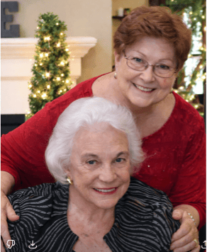 Two smiling older women pose together indoors. One, with short white hair and a zebra-striped jacket, is seated, while the other stands behind her in a red top, hugging her shoulders. A decorated tree twinkles in the background at The Citizen.
