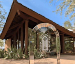 A glass National Preservation Award trophy is displayed in front of a mid-century modern, triangular-roofed building, celebrating historic preservation amid lush greenery and a bright blue sky.