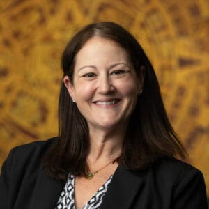 A woman with long brown hair, wearing a black blazer and patterned blouse, smiles at the camera. The background is blurred with warm golden tones and circular patterns.