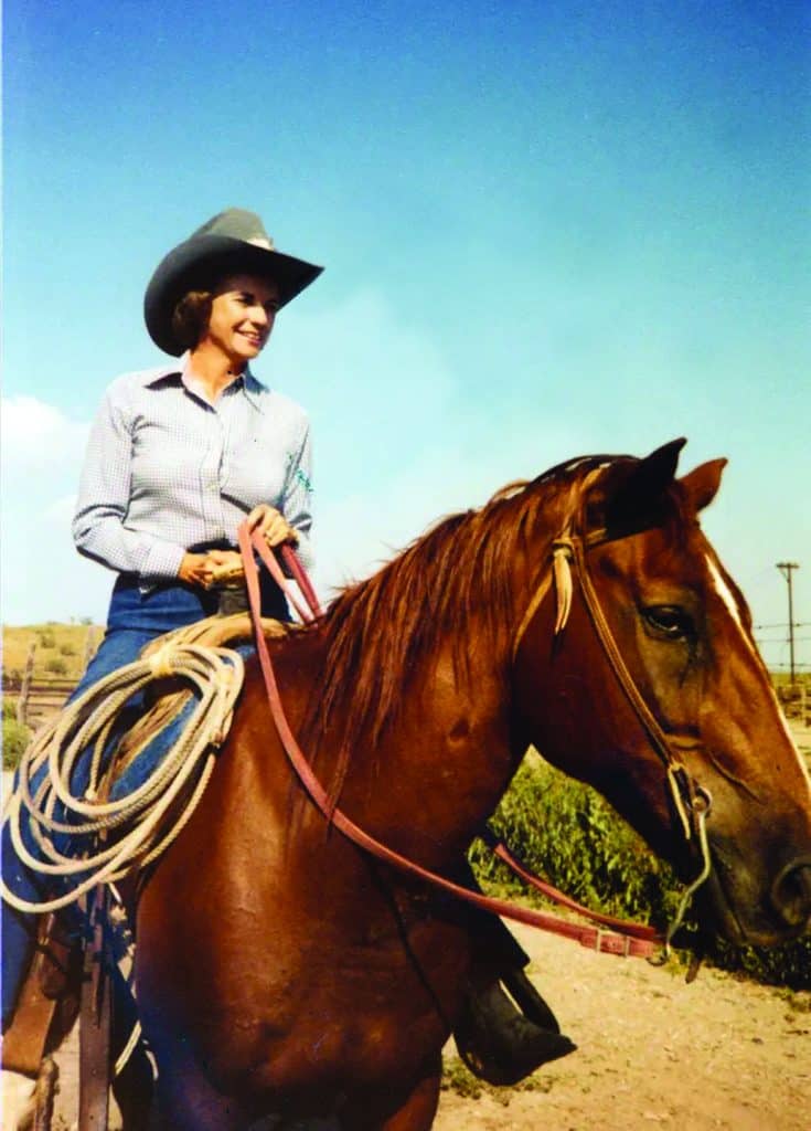 A woman wearing a cowboy hat and checkered shirt rides a brown horse outdoors on a sunny day, ready to build her path through the open land. Coiled rope hangs by her side, with blue sky and distant hills in the background.