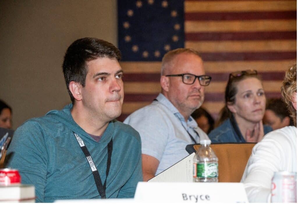 A group of people sit attentively at a conference or meeting, wearing name badges, as an old American flag—symbolizing the Constitution—hangs in the background. A water bottle and a name tag labeled “Bryce” are on the table in front.