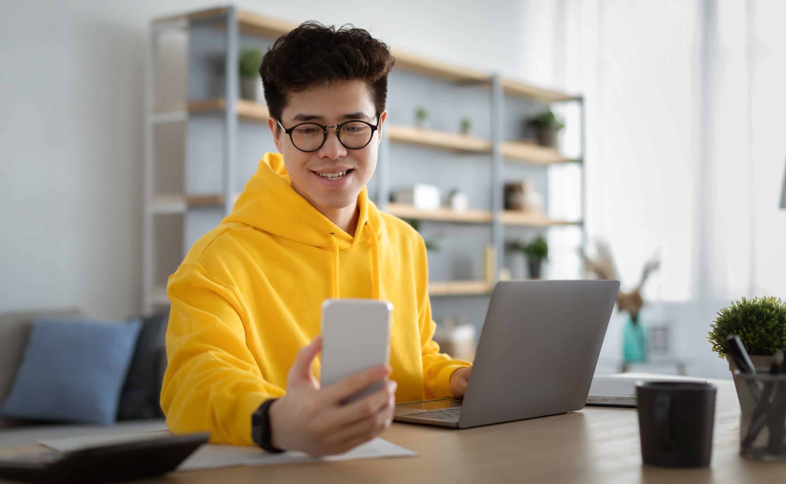 Young person wearing a yellow hoodie and glasses sits at a desk with a laptop, smiling while holding a smartphone for a selfie or video call, showcasing their Civics for Life enthusiasm in a bright, modern home office.