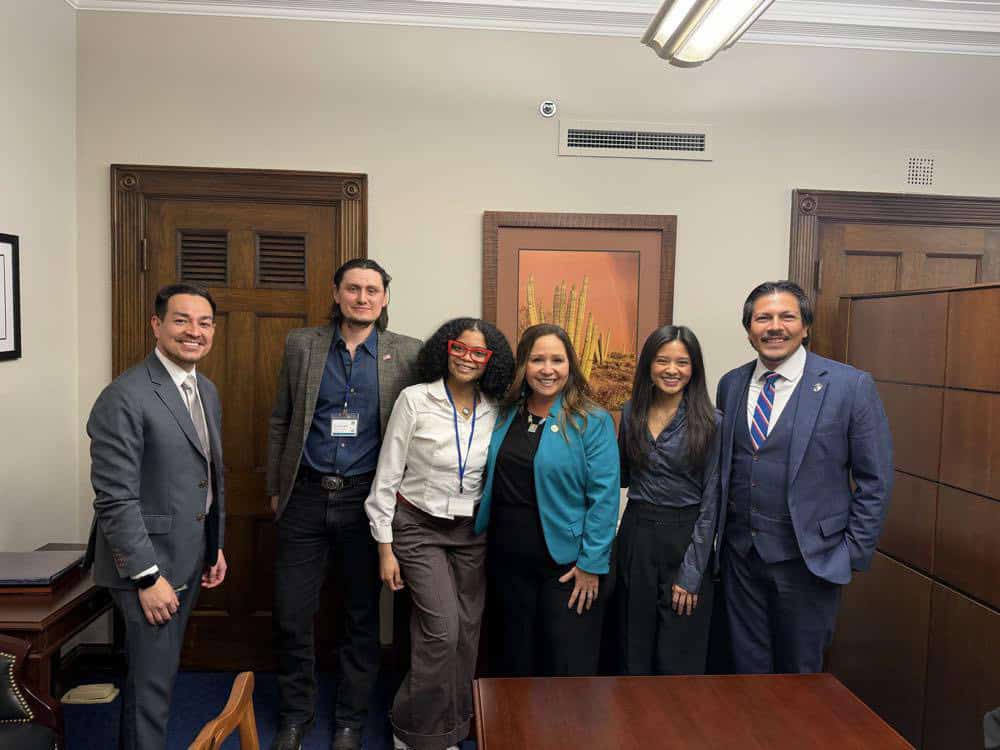 Six people in business attire stand together and smile in an office with wooden doors, a framed picture of cacti on the wall, and a desk in the foreground—capturing the spirit of Capital Connections.