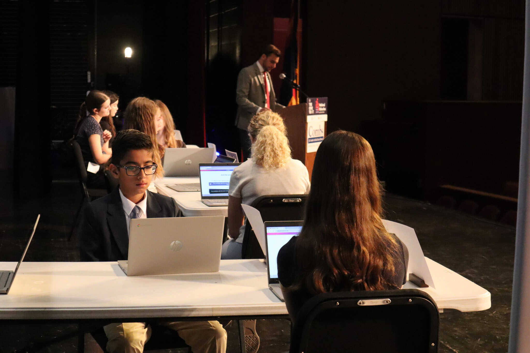 Several students in formal attire sit at tables with laptops on a stage, facing away from the audience. A man stands at a podium in the background, speaking into a microphone.