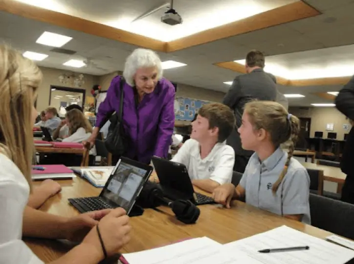 An elderly woman in a purple jacket discusses self governance with three students seated at a table using laptops in a busy classroom or library setting. Other students and adults are visible in the background.