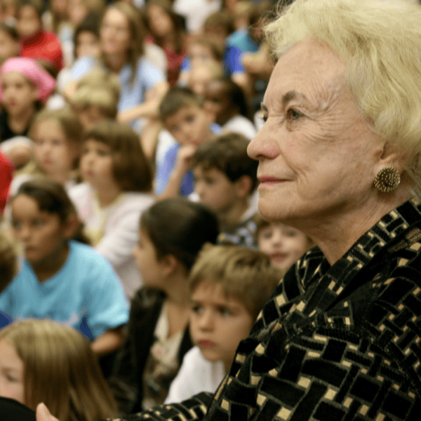 An older woman with short blonde hair wearing a patterned jacket sits among a crowd of children. The children are seated and appear to be watching a presentation or event. The focus is on the woman's profile and attentive expression.