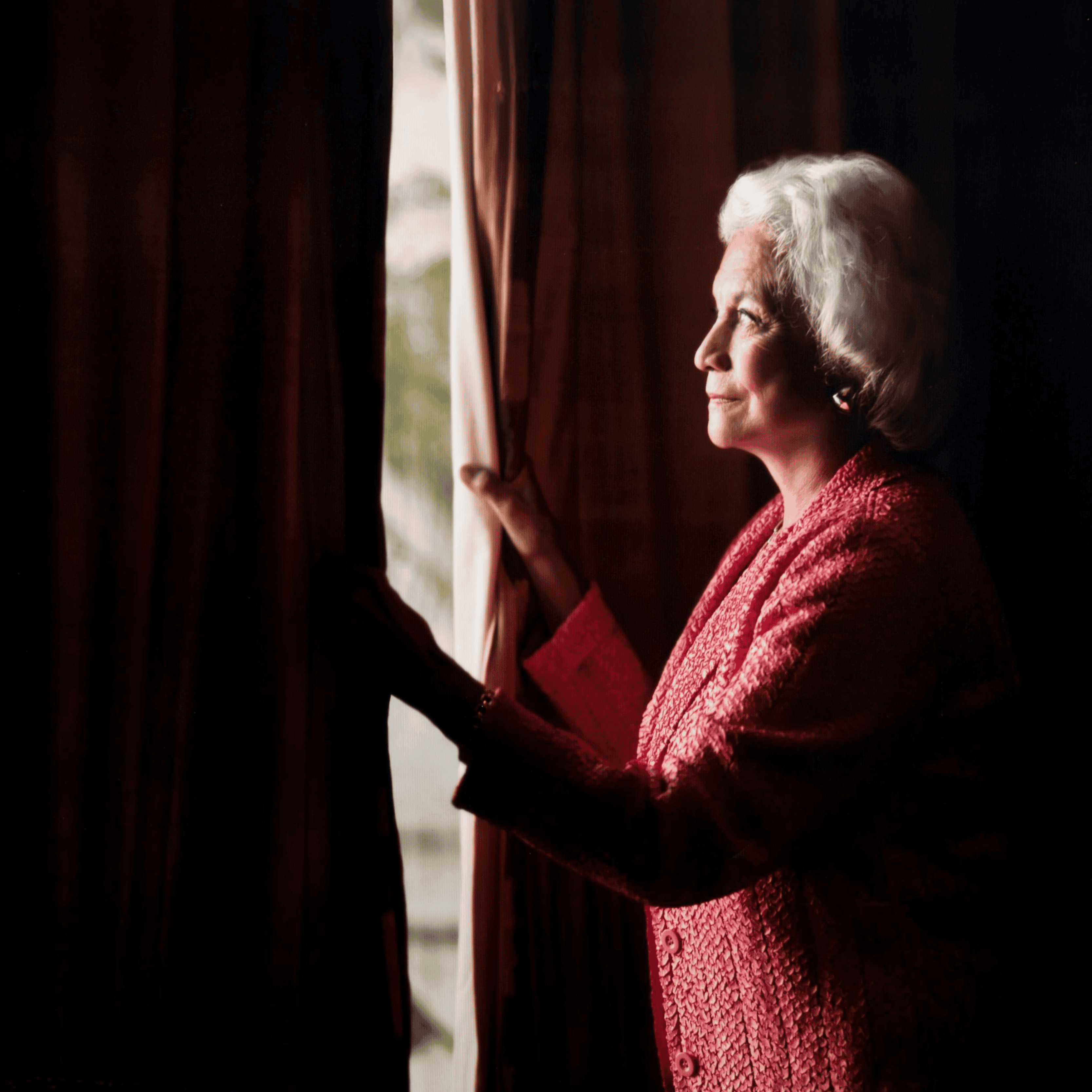 An elderly woman with short white hair stands by a window, holding the curtains open slightly, gazing outside. She is wearing a textured pink jacket. The light creates a soft glow around her.