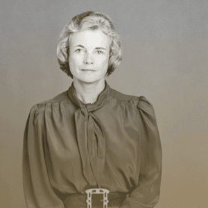 A woman with short, wavy gray hair wears a long-sleeved blouse with a necktie and a wide belt, facing the camera with a neutral expression. The simple, gradient background evokes the professional setting of Youth Programs.