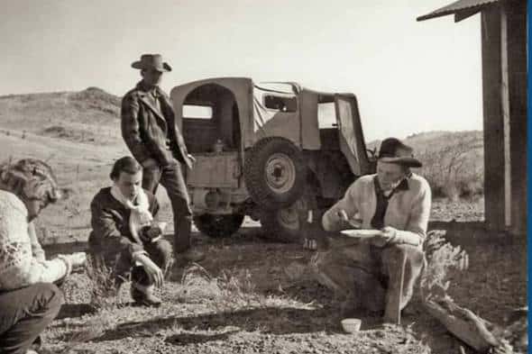 Four people in cowboy hats eat outdoors near a Jeep and a small shed in a rural, hilly landscape. The black-and-white scene hints at camaraderie and justice under open skies as they share a meal amid the rugged terrain.
