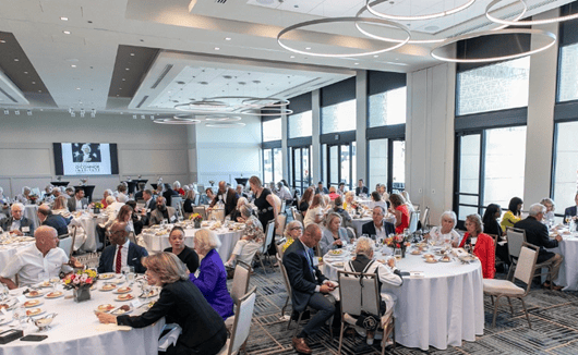 A large group, including Dr. Summers, sits at round tables in a spacious, modern banquet hall with big windows, eating and conversing during a formal event. Flowers and plates of food are on the tables.