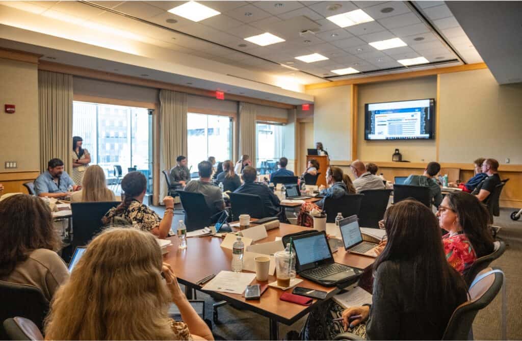 A group of people sit at tables in a conference room, watching a presentation on the Constitution displayed on a large screen. Some are using laptops, while others take notes or listen attentively.