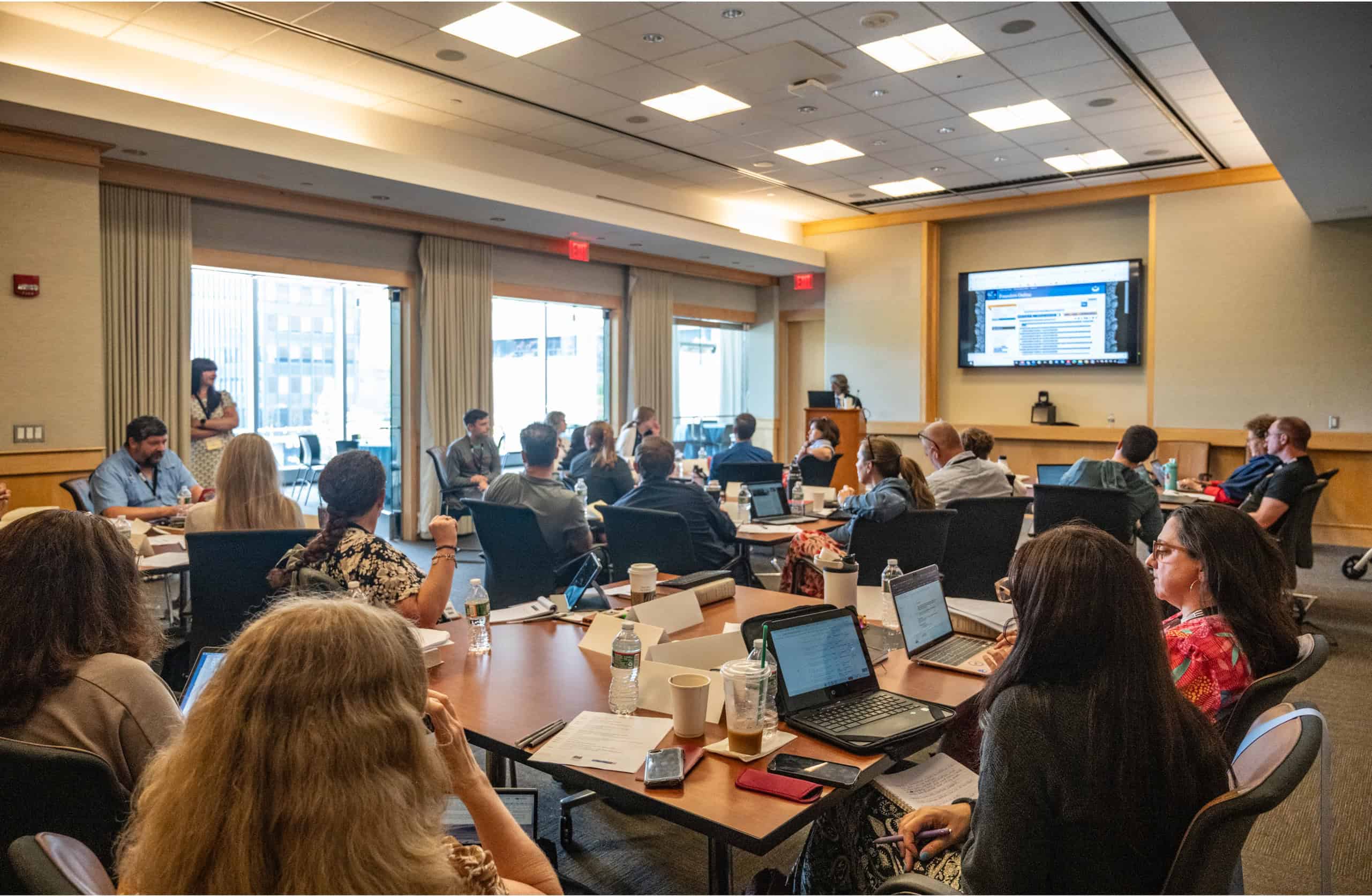 A group of people sit at tables in a conference room, watching a presentation on the Constitution displayed on a large screen. Some are using laptops, while others take notes or listen attentively.