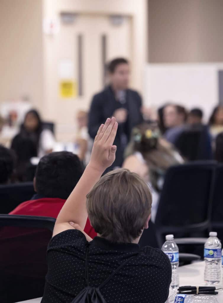 A student with short hair raises their hand in a classroom or seminar at The Citizen, facing a blurred speaker at the front. Other attendees sit nearby, and water bottles are visible on the table.