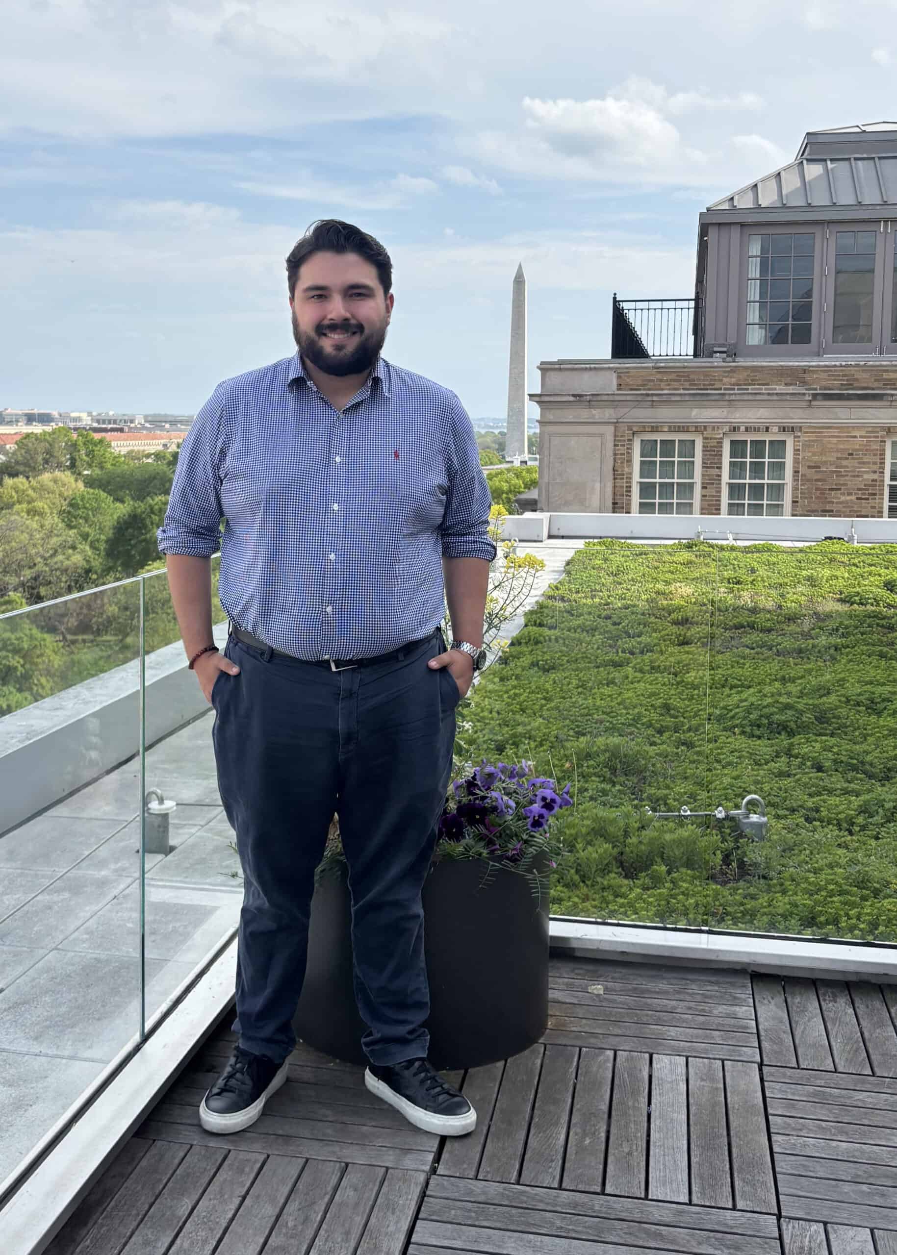 A man stands on a rooftop deck with a green roof, glass railing, and potted flowers. The Washington Monument is visible in the background under a partly cloudy sky.