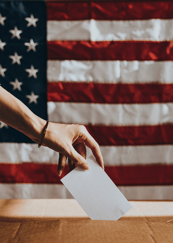 A person’s hand places a white ballot into a box, with a large American flag in the background, highlighting the importance of Capital Connections in shaping democracy.
