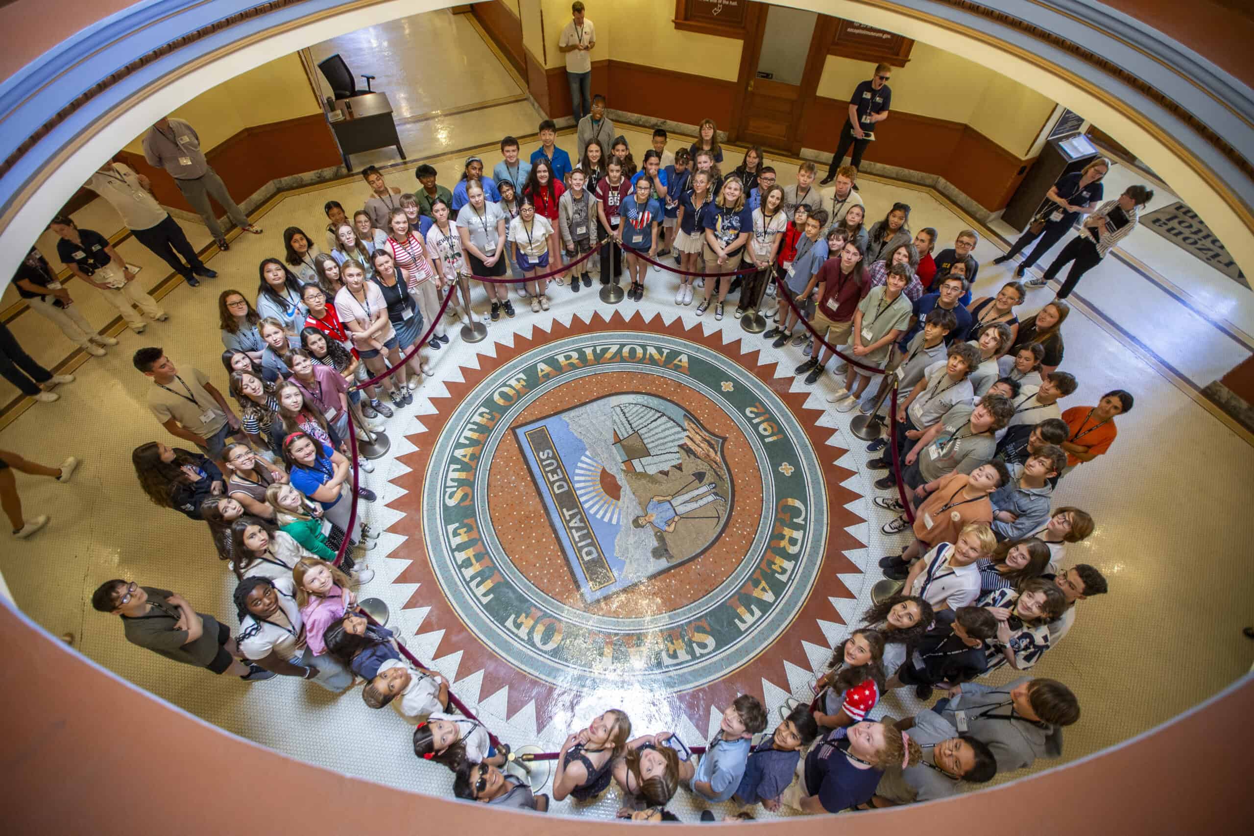 A large group of people, mostly children, stand in a circle around the Great Seal of the State of Arizona on the floor inside a government building, as each citizen looks up at the camera.