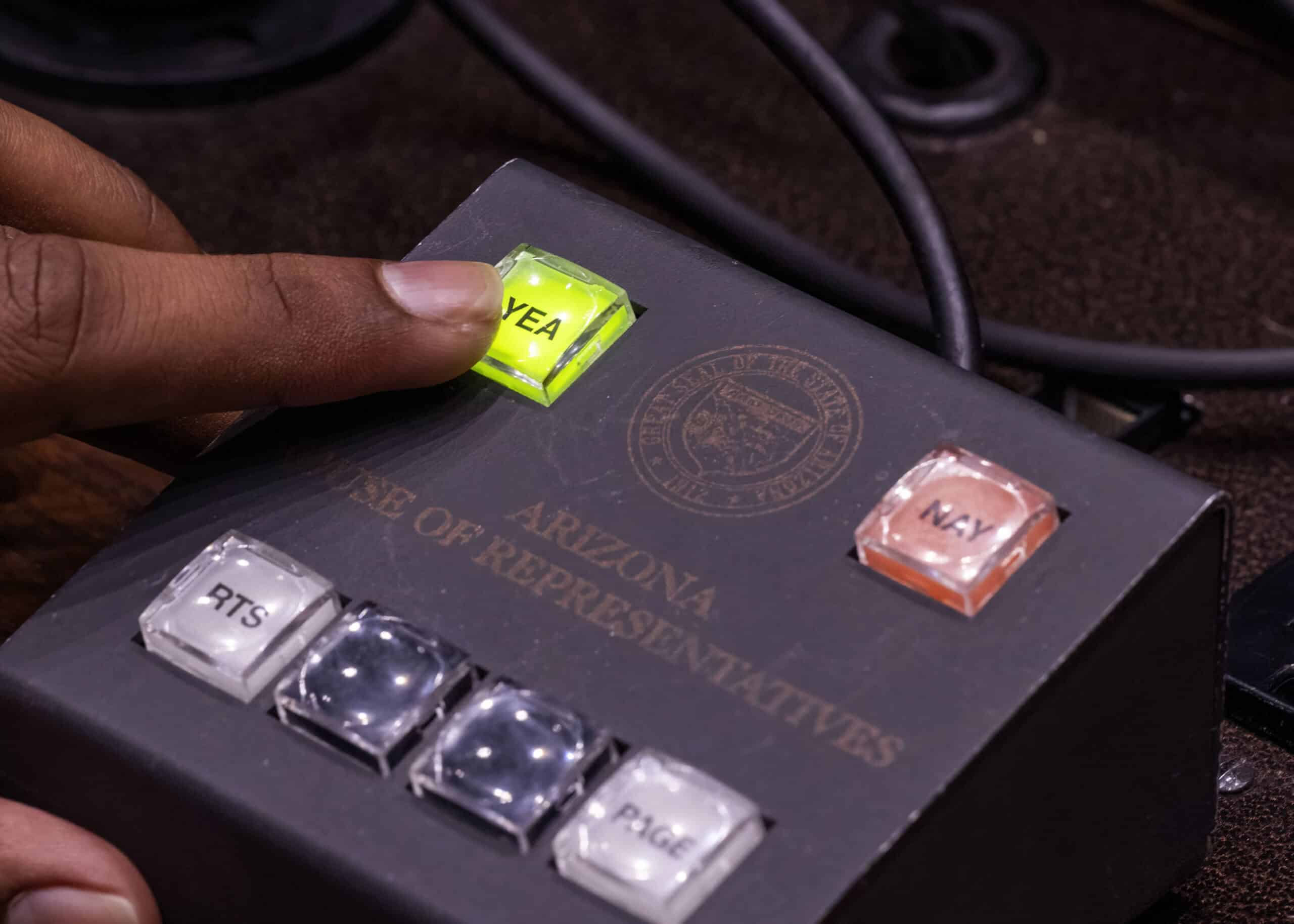 A hand presses the glowing green "YEA" button on a voting device labeled "Arizona House of Representatives," while The Citizen observes other buttons marked "NAY," "PTS," and "PAGE.