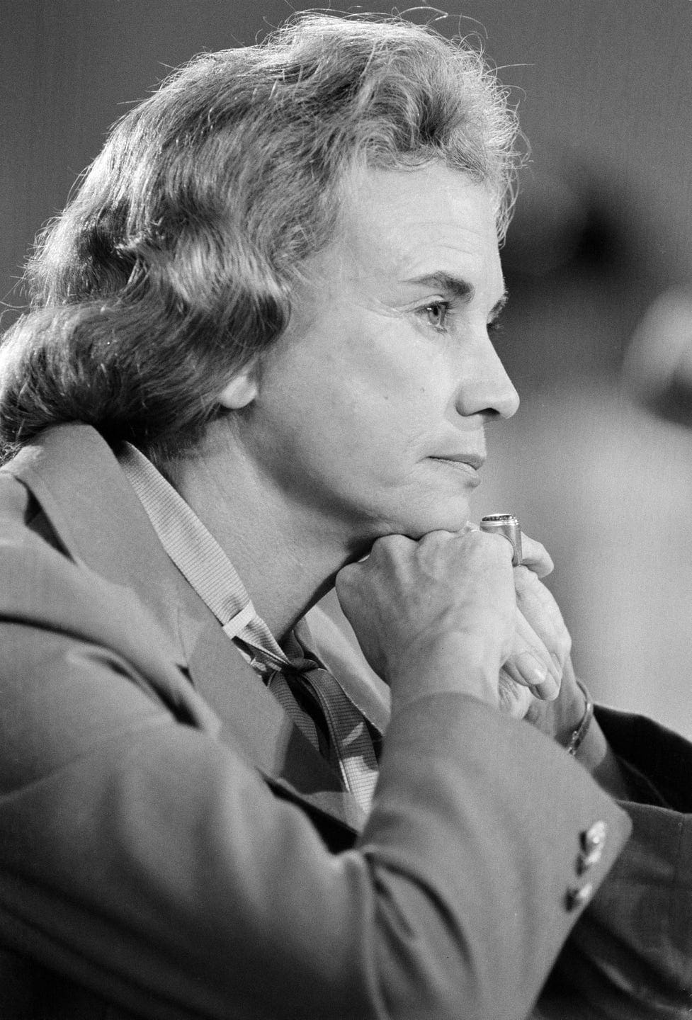 A person with shoulder-length hair rests their chin on folded hands while looking thoughtfully to the side. They are wearing a suit, and the image is in black and white.