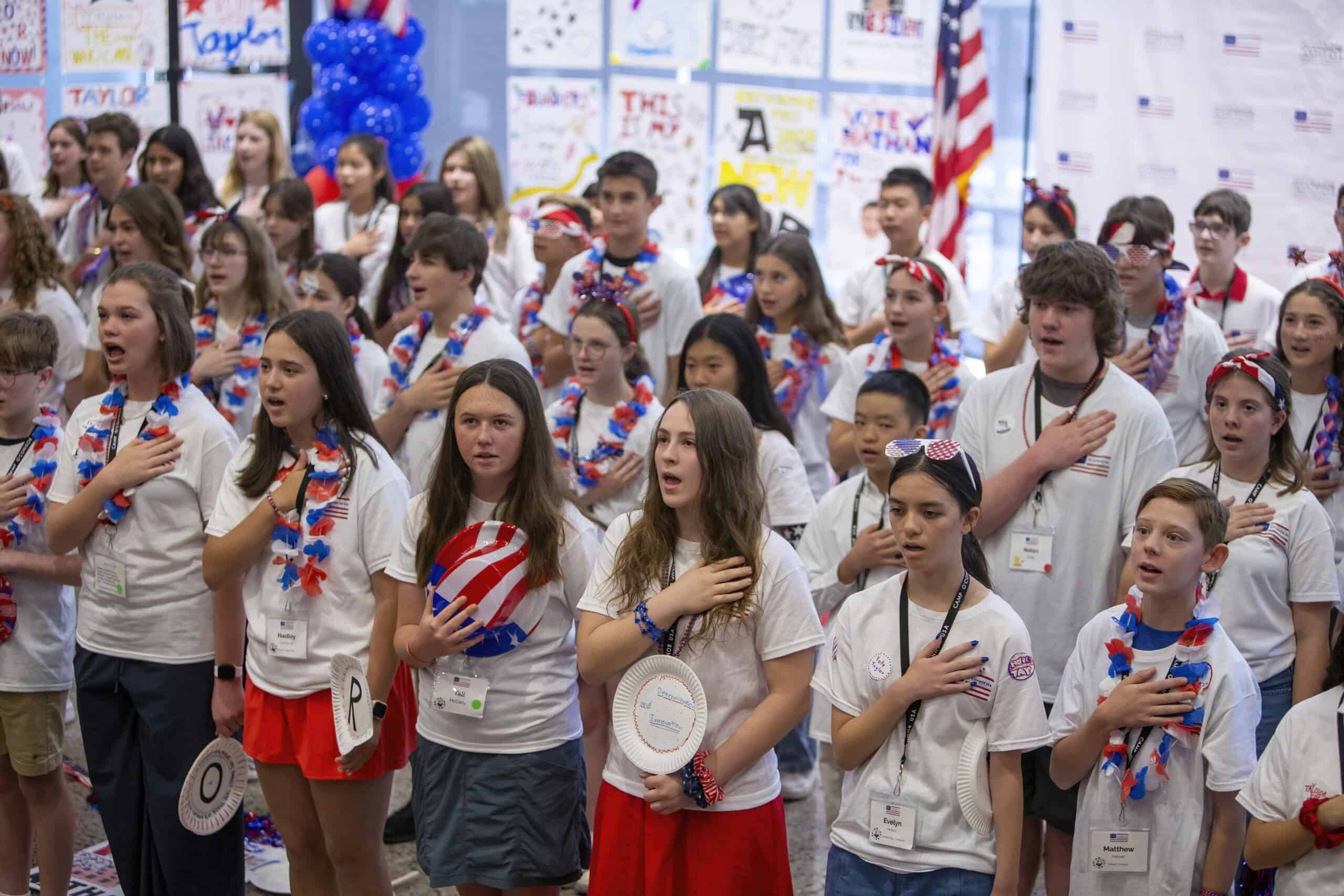 A large group of young citizens wearing red, white, and blue clothing and accessories stand with their right hands over their hearts, appearing to recite the Pledge of Allegiance at a patriotic event. Patriotic posters and an American flag are visible.