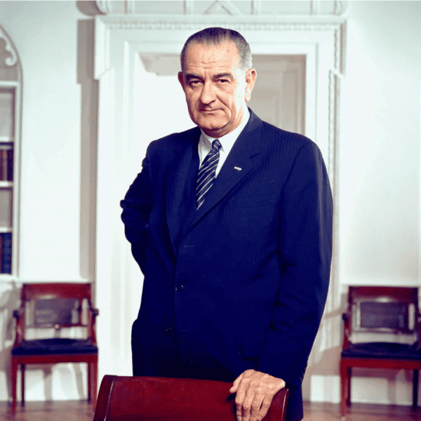 An older man in a suit stands confidently in an elegant room with white walls and ornate details, reminiscent of a grand history. Wooden chairs form the backdrop as he rests one hand on a chair, exuding poise as if poised for an important dinner. The room is softly lit.